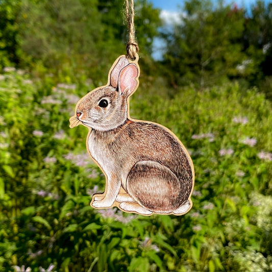 Wooden rabbit ornament hanging against a natural background with greenery and flowers.