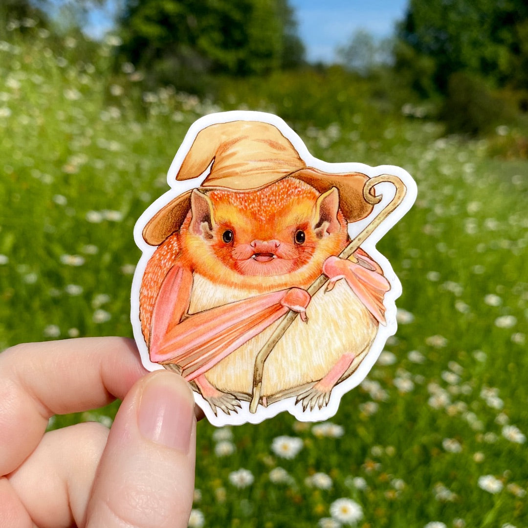 A hand holding a sticker of a round eastern red bat carrying a twig staff and with a brown wizard hat.