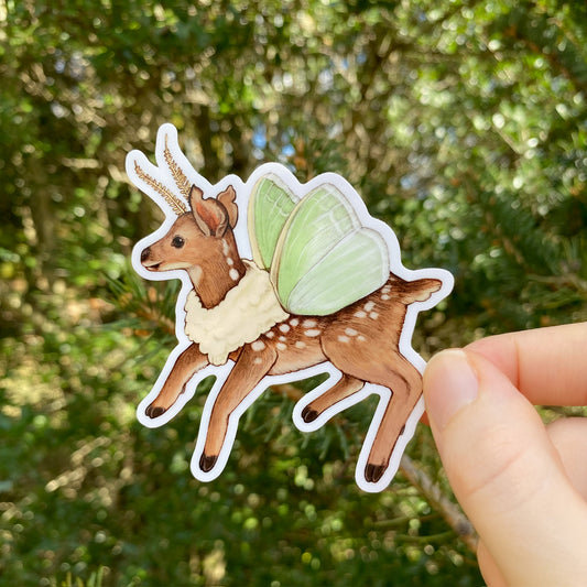 A hand holding a sticker of a white-tailed deer fawn with emerald moth wings. 