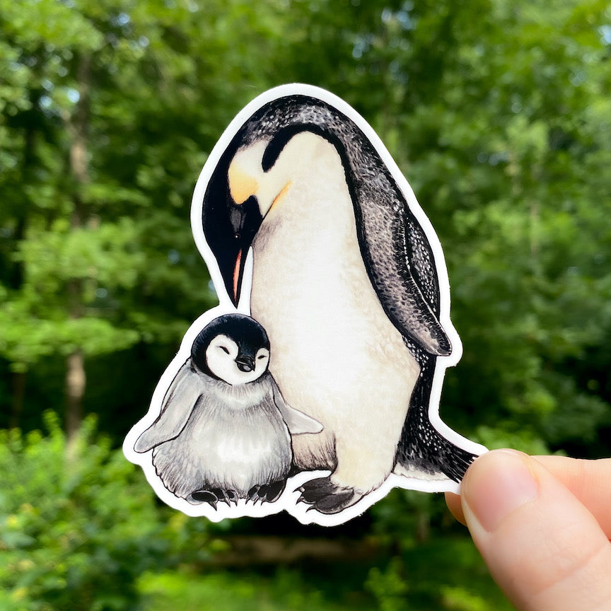 A hand holding a sticker featuring an illustration of an emperor penguin and chick