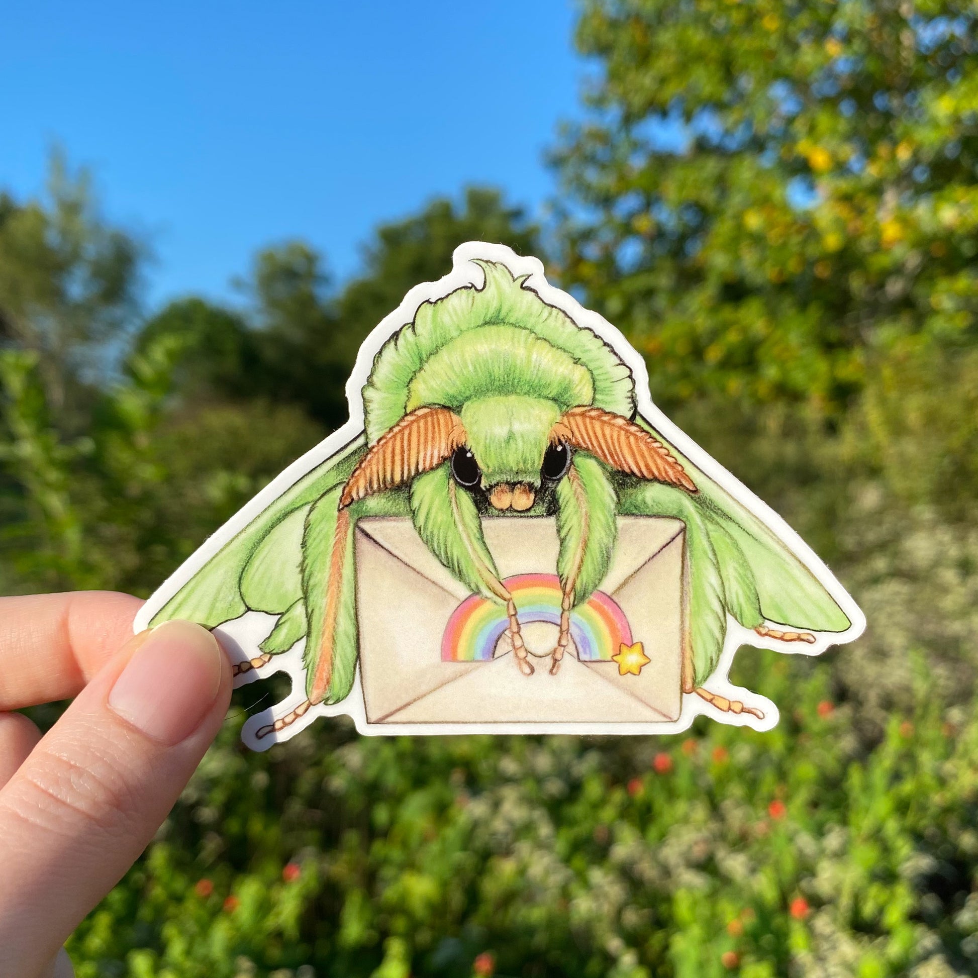 A hand holding a weatherproof vinyl sticker of a green moth holding a letter with a rainbow sticker.