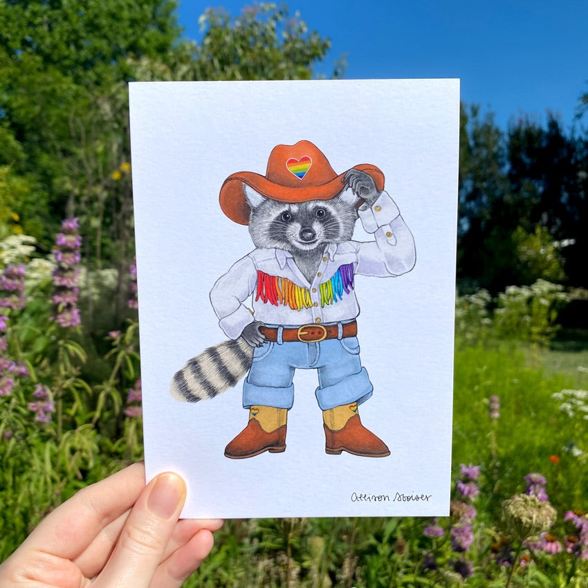 A hand holding a print of a raccoon dressed up in a rainbow cowboy outfit.