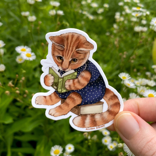Sticker of an orange cat reading a book held in front of a natural background with flowers.