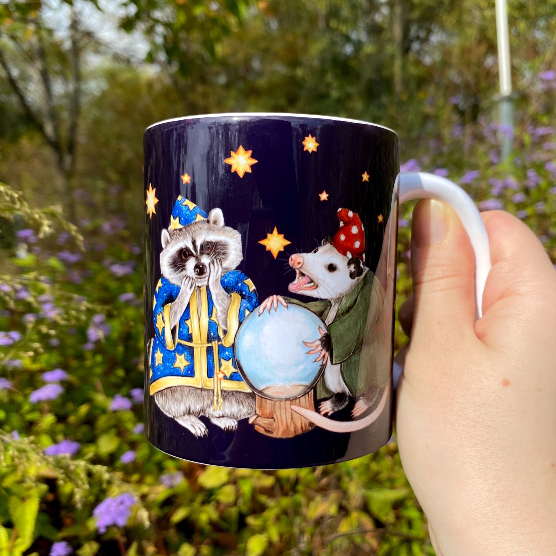 Mug with a wizard raccoon and opossum looking into a crystal ball, held by a hand in a natural setting.
