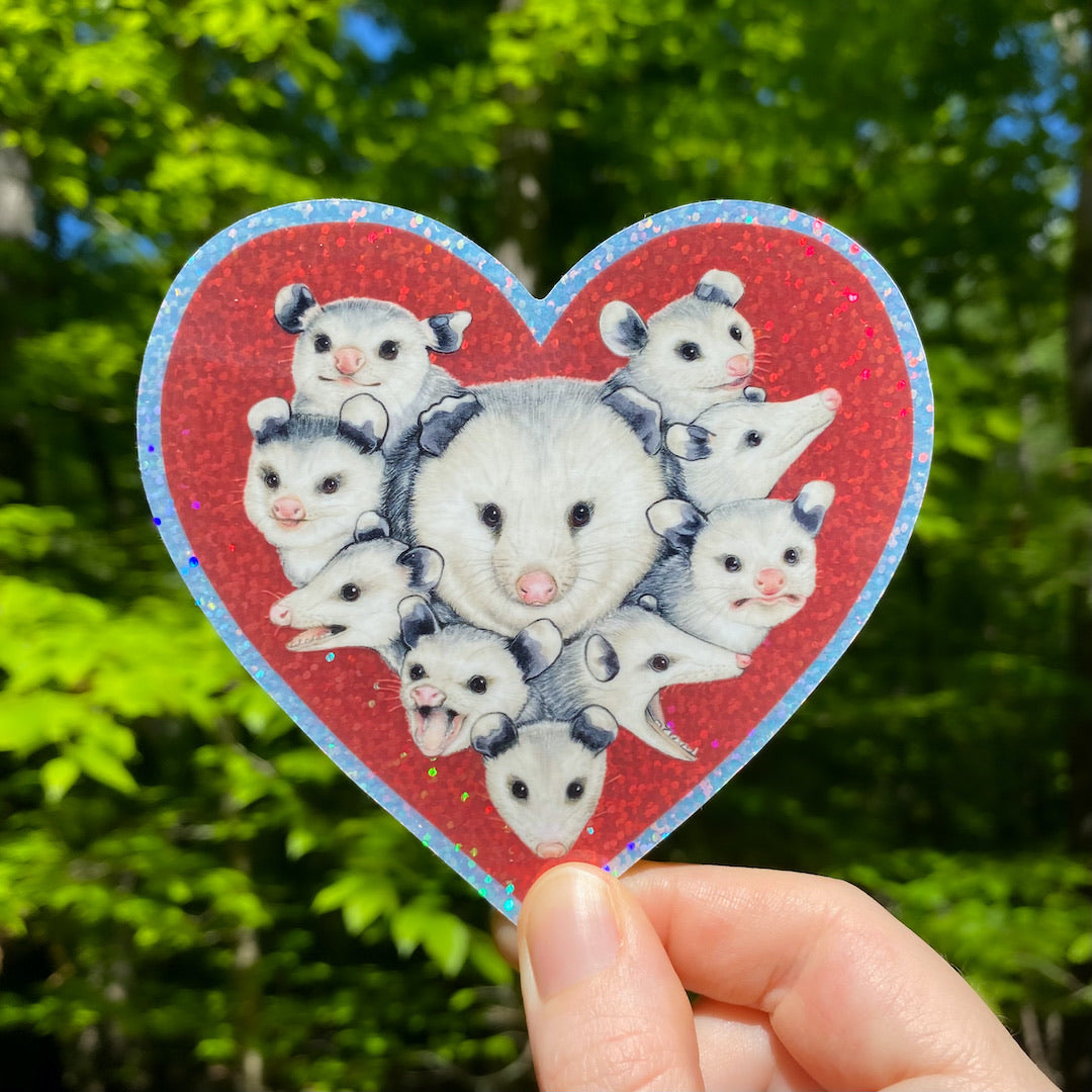 A hand holding a weatherproof vinyl sticker of mom opossum surrounded with baby opossums in a red sparkly heart.