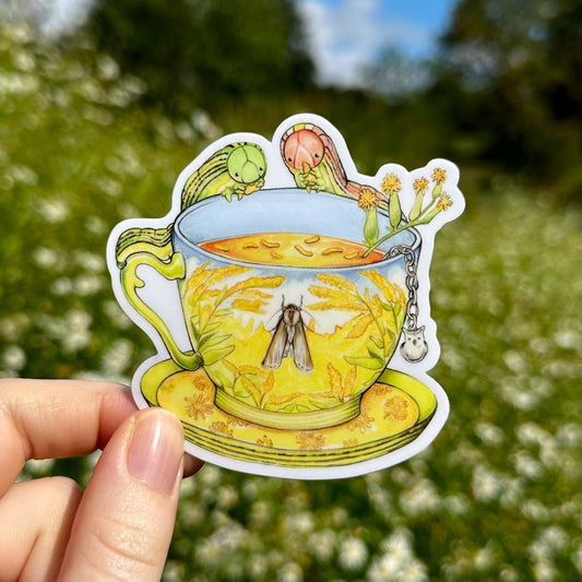 My hand holding a sticker of a teacup decorated with golden rod flowers on a blue sunny background with green caterpillars on the rim.