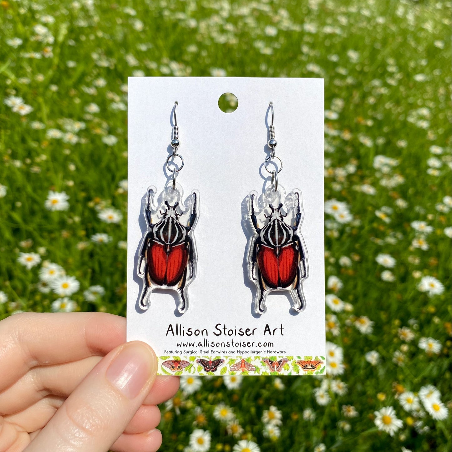 A hand holding a pair of clear acrylic earrings of goliath beetles.