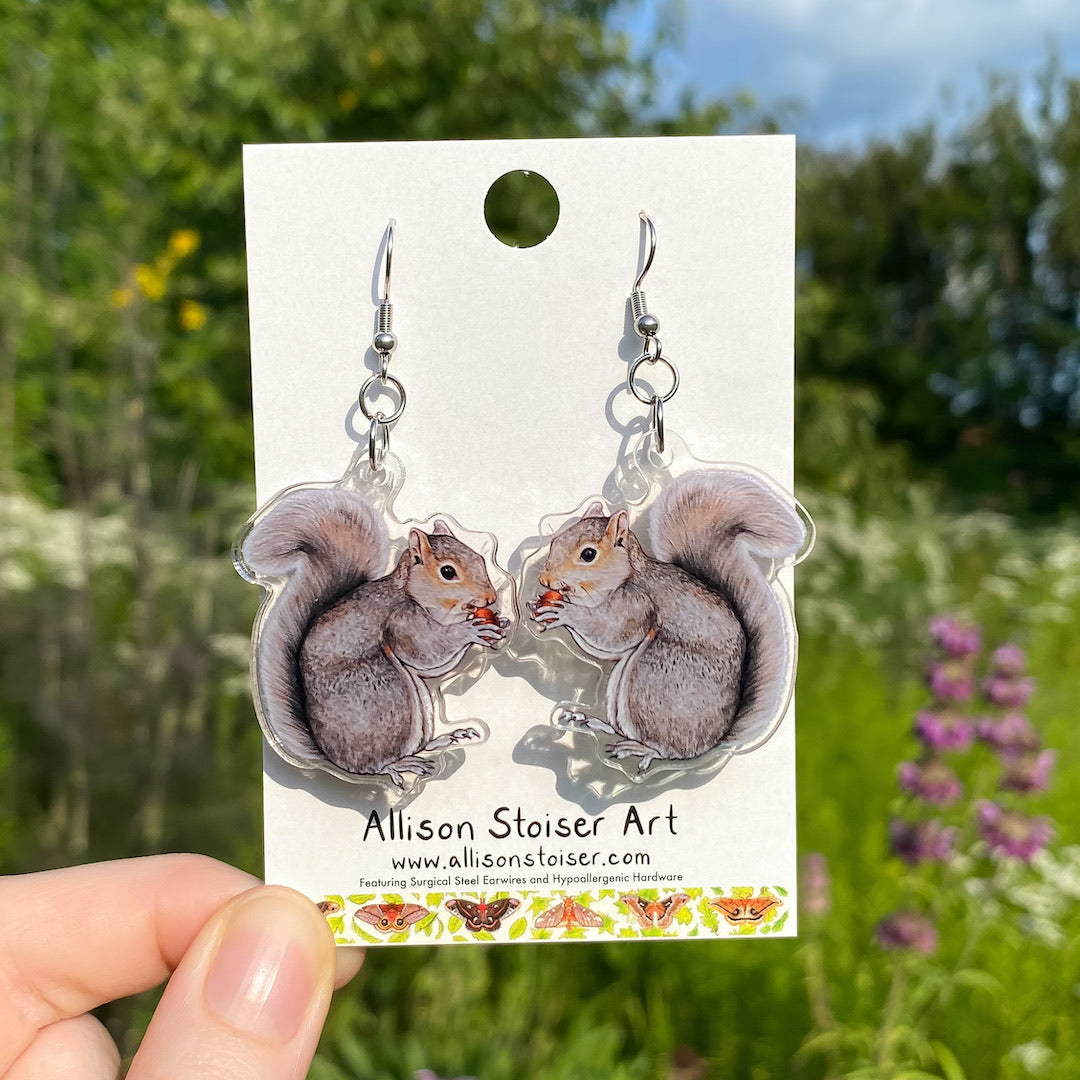 A hand holding a pair of acrylic earrings of gray squirrels holding acorns.