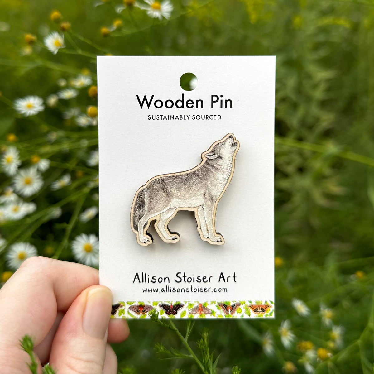 A wooden pin of a howling wolf, mounted on a white backing card held by a hand against a blurred background of green foliage and asters.