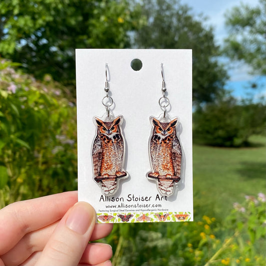 A hand holding a pair of acrylic earrings of great horned owls.