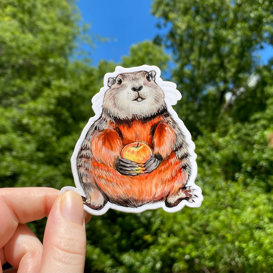 A hand holding a sticker of a groundhog holding an apple