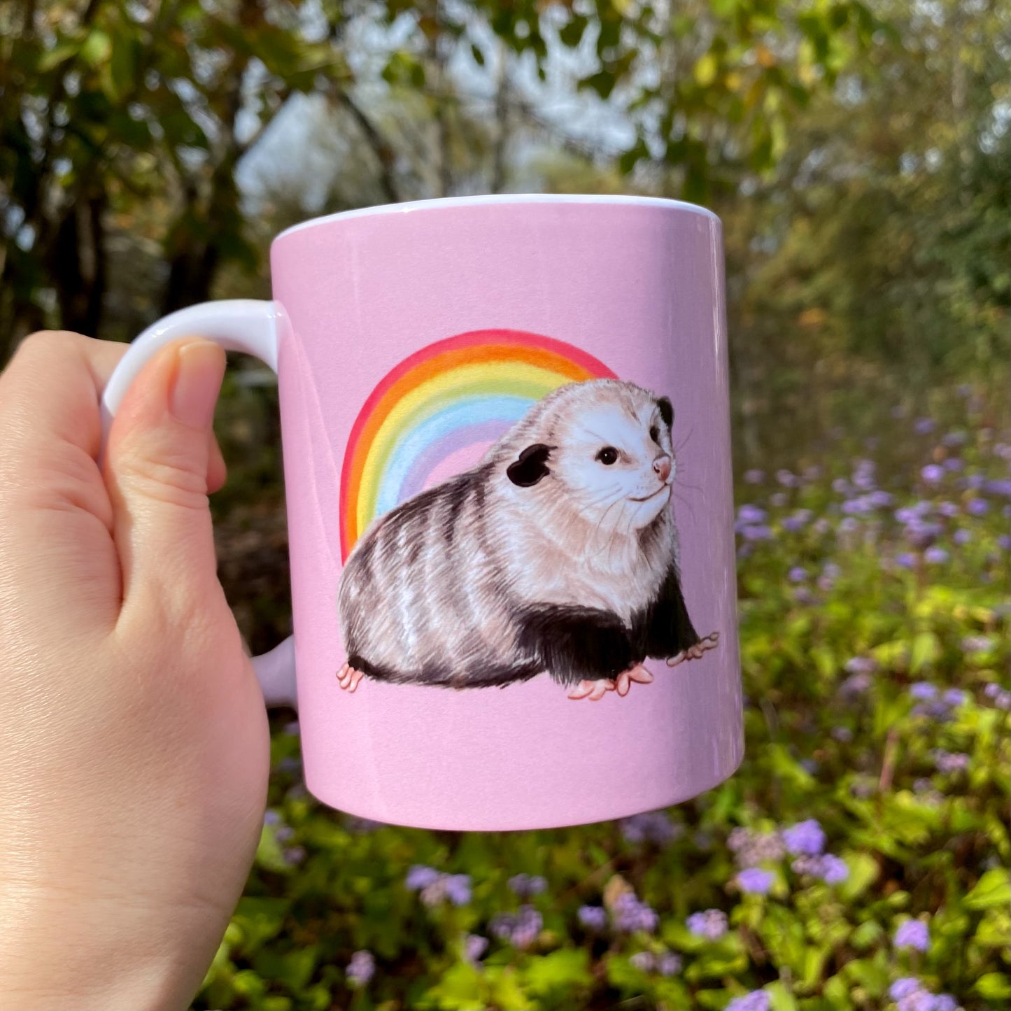 Pink mug with an opossum and rainbow illustration held by a hand outdoors.