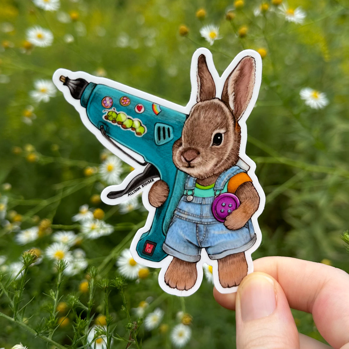 A die-cut sticker featuring a brown rabbit in blue overalls holding a teal hot glue gun and button, shown being held by a hand against a blurred outdoor background of green plants and asters.