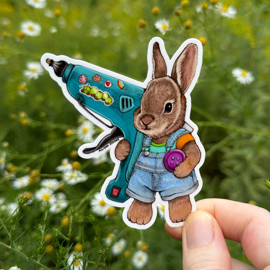 A die-cut sticker featuring a brown rabbit in blue overalls holding a teal hot glue gun and button, shown being held by a hand against a blurred outdoor background of green plants and asters.