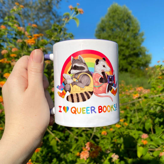 Mug with raccoon and opossum illustration and 'I ❤️ QUEER BOOKS!' text, held outdoors.