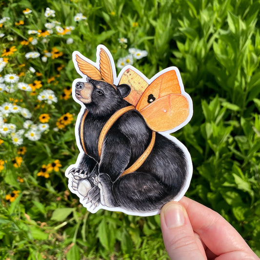 A hand holding a weatherproof vinyl sticker of a black bear dressed up as an io moth.