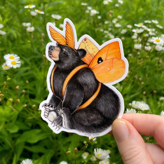 Sticker of a bear with io moth wings held in front of a natural background with flowers.