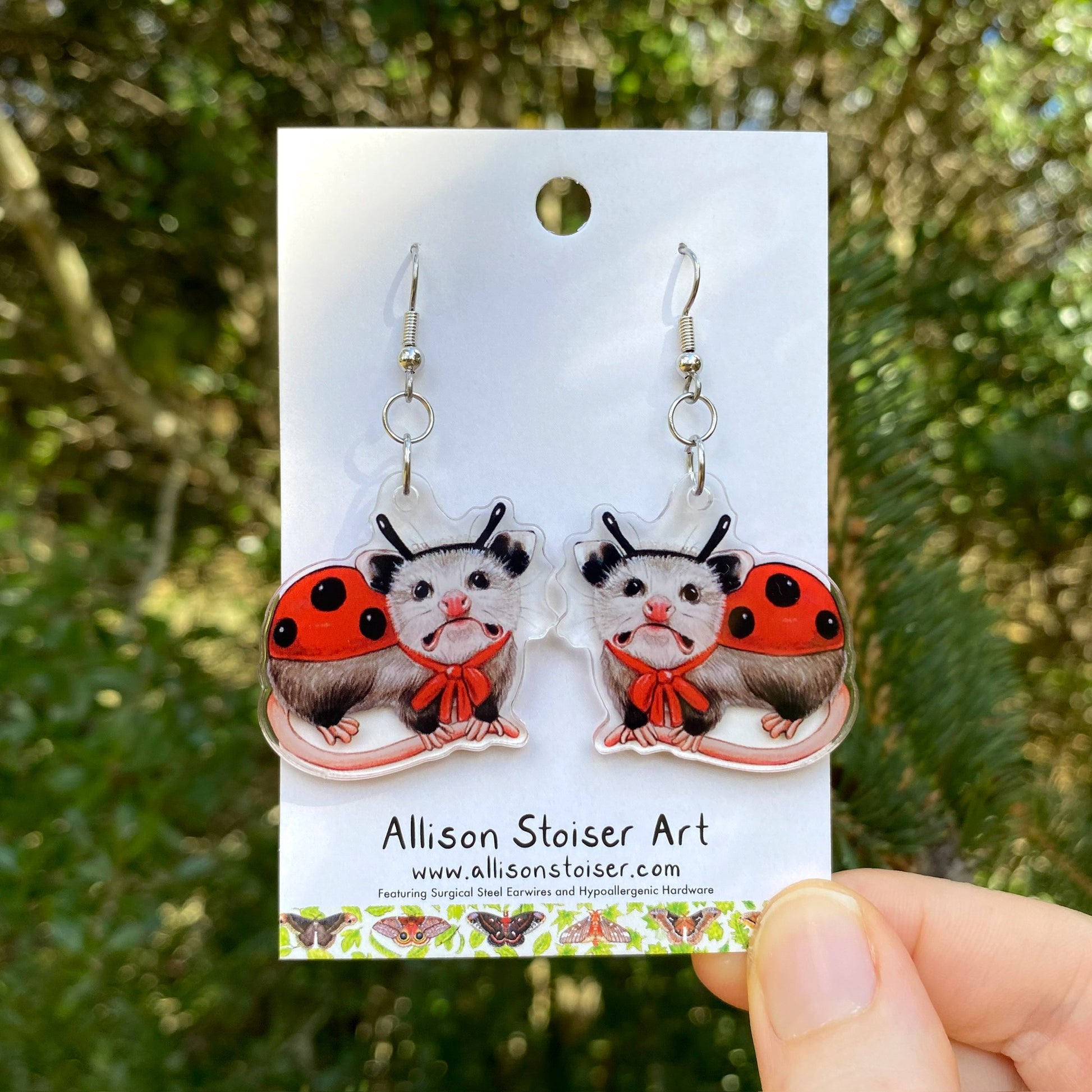 A hand holding a pair of ladybug opossum acrylic earrings