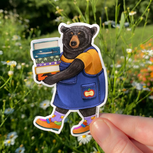 Sticker of a bear holding books, held by a hand against a natural background with flowers and greenery.