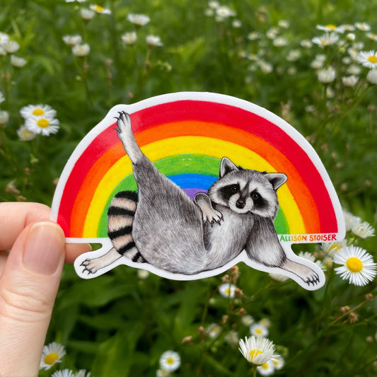 Sticker of a raccoon on a rainbow with a hand holding it, against a natural background with flowers.