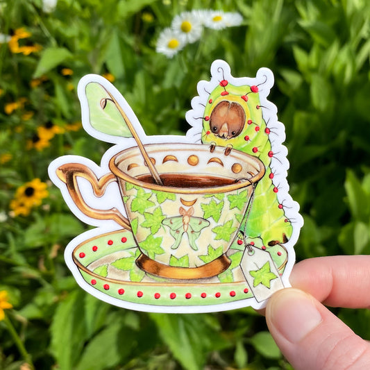 A hand holding a weatherproof vinyl sticker of a delicate teacup with sweetgum leaf designs with a luna moth caterpillar sitting at the rim..