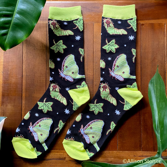 A pair of black and green socks on a wood table with luna moths and caterpillars. 