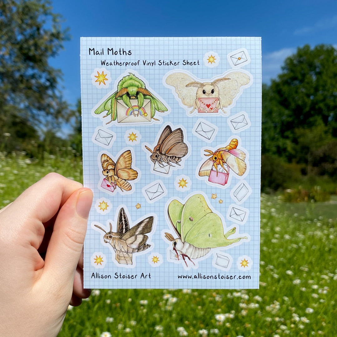 A hand holding a sticker sheet that reads "Mail Moths Weatherproof Vinyl Sticker Sheet" along the top and "Allison Stoiser Art" and "allisonstoiser.com" along the bottom. The sticker sheet is decorated with tiny stickers of envelopes and stars, and seven moths carrying letters. The moths are all different species, some green, some brown, some orange, and some beige.