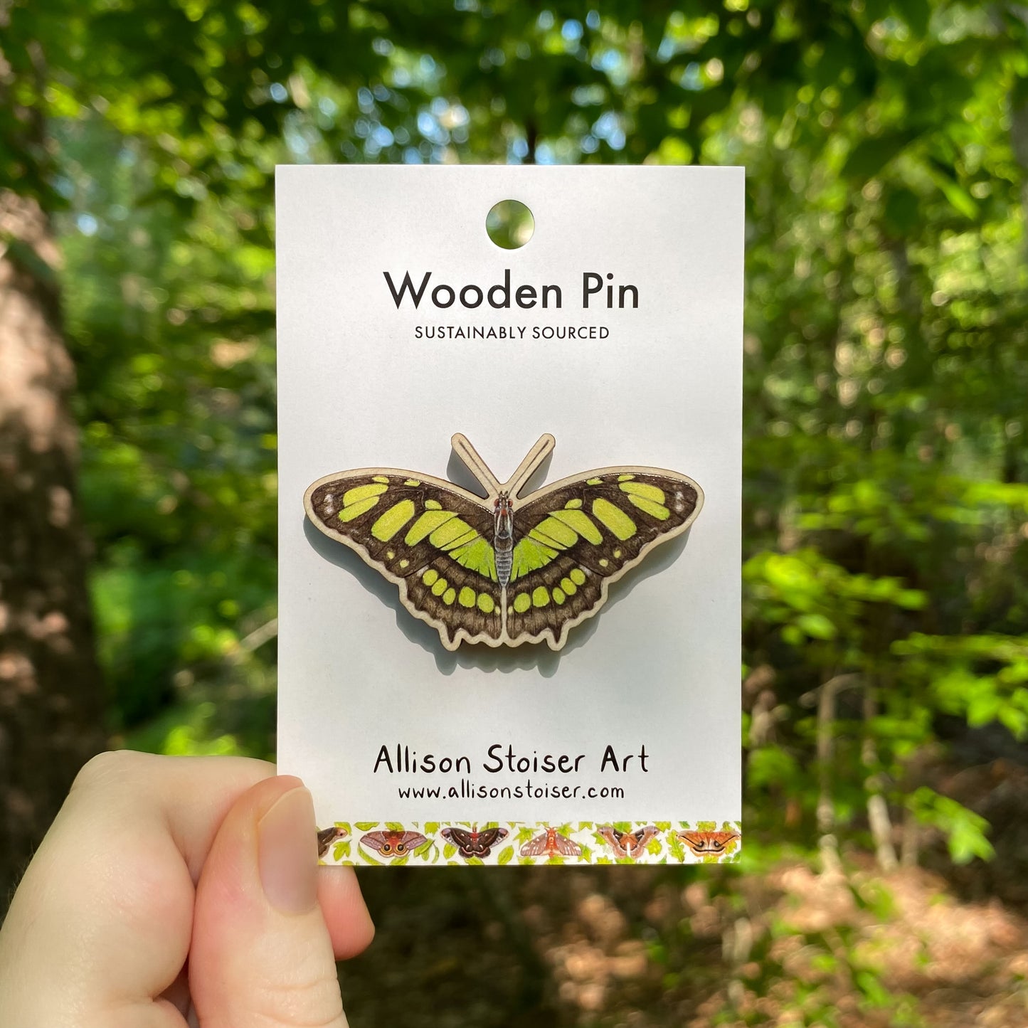 A wooden pin of a green malachite butterfly on packaging that reads Wooden Pin, Sustainably Sourced.