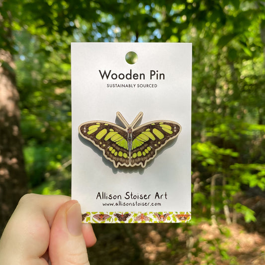 A wooden pin of a green malachite butterfly on packaging that reads Wooden Pin, Sustainably Sourced.