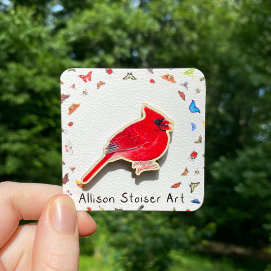 A hand holds a sustainably sourced wooden pin featuring an illustration of a male northern cardinal