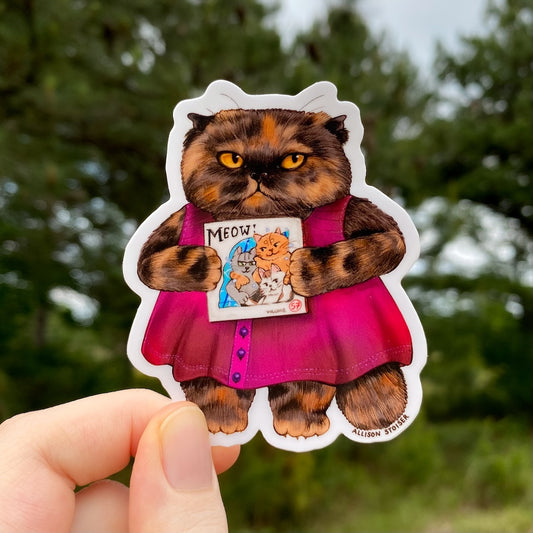 Sticker of a tortoiseshell Persian cat in a pink dress held by a hand with a blurred green background