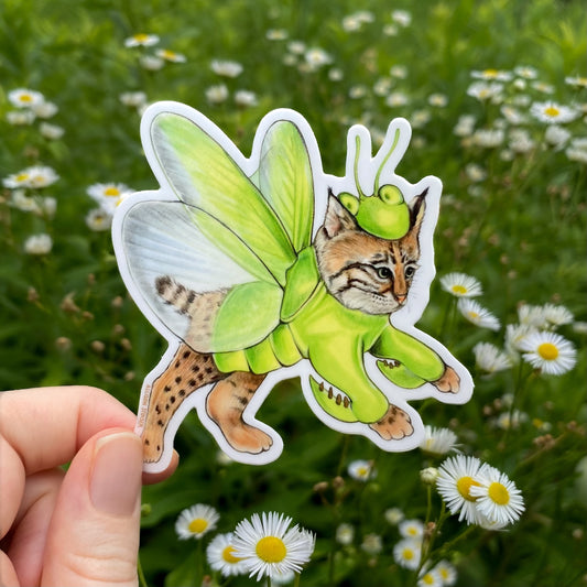 Sticker of a cat in a green praying mantis costume held in front of a grassy background with flowers.