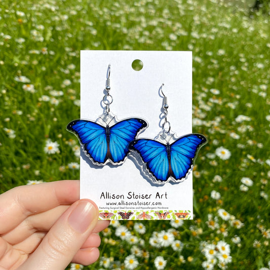 A hand holding a pair of clear acrylic earrings of blue morpho butterflies.