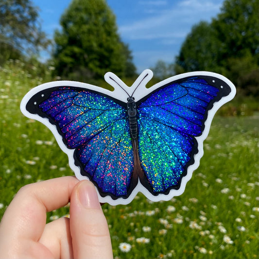 A hand holding a glittery sticker of a blue menelaus morpho.