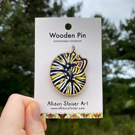 Wooden pin of a monarch caterpillar on a card with 'Allison Stoiser Art' branding against a blurred natural background