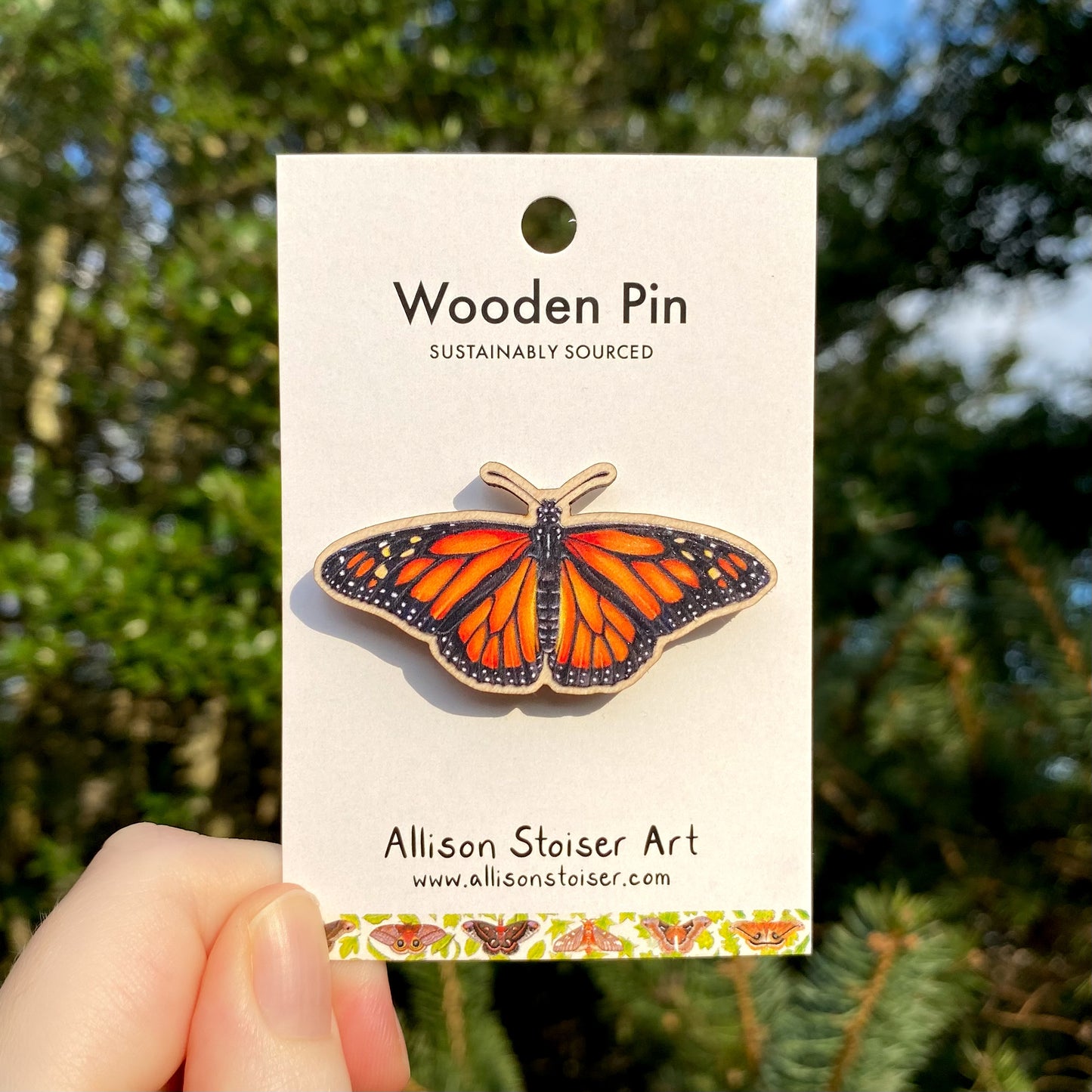 A wooden pin with a monarch butterfly design, held in front of a blurred natural background.