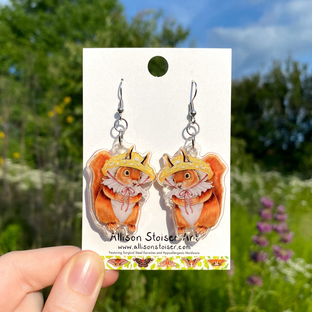 A hand holding a pair of acrylic earrings with red squirrels in mushroom hats.