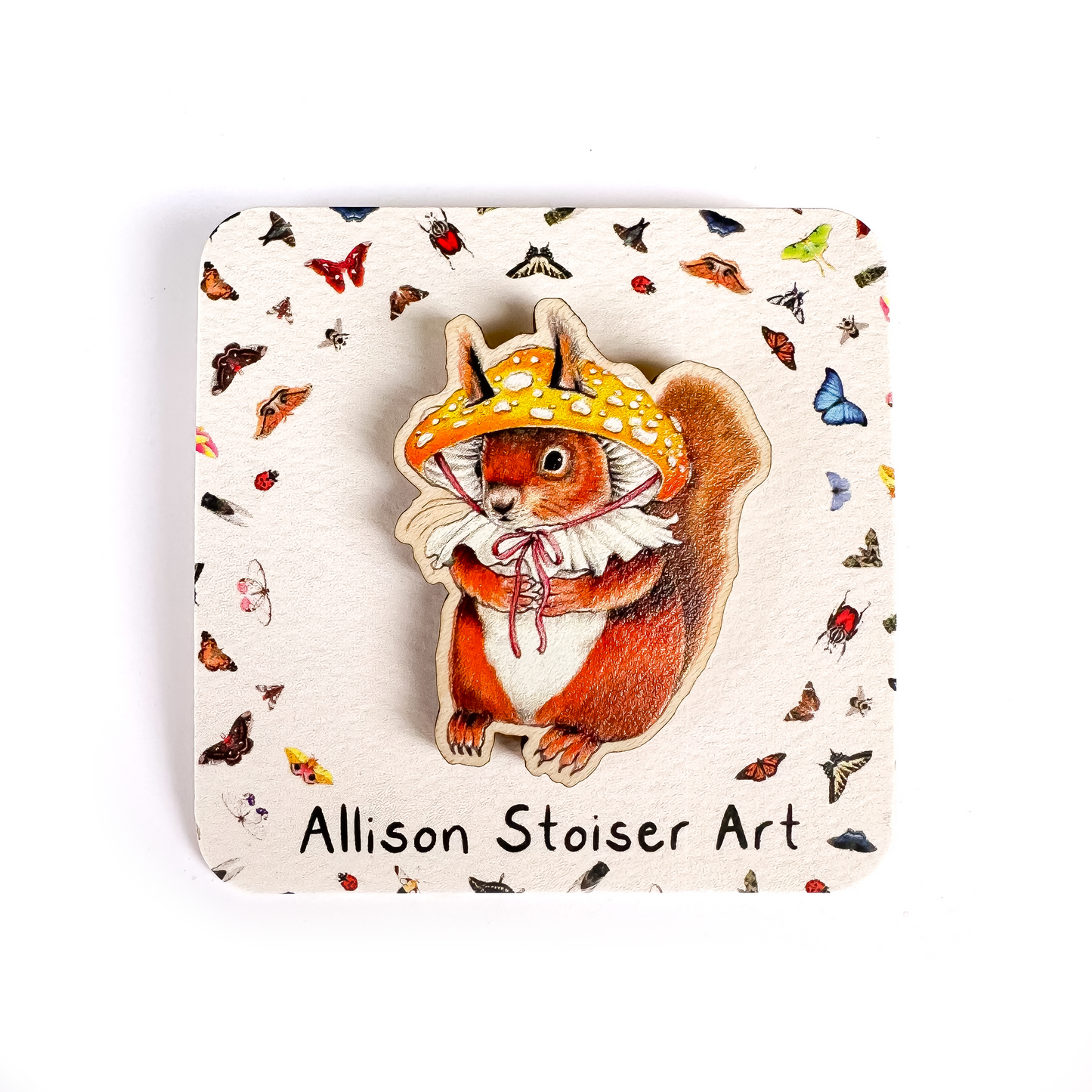 A wooden pin of a red squirrel wearing a yellow patches mushroom hat on a white background.