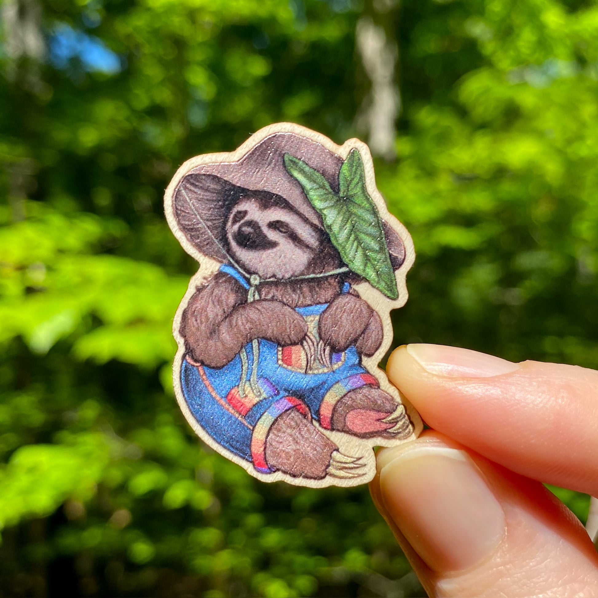 A hand holding a wooden pin of a sloth in overalls wearing a mushroom hat.