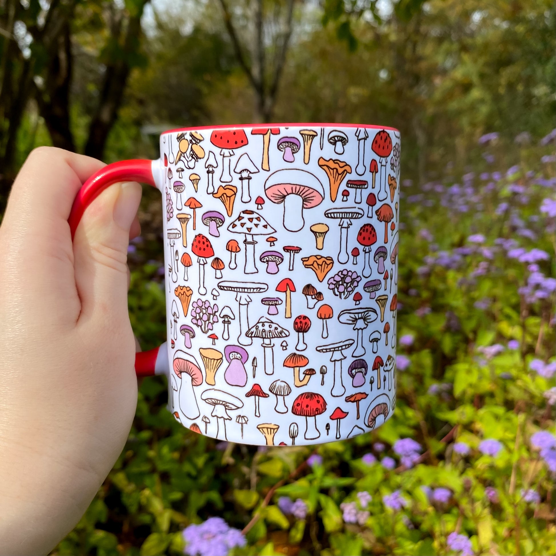 Mug with mushroom pattern held by a hand in a natural setting with flowers and trees.