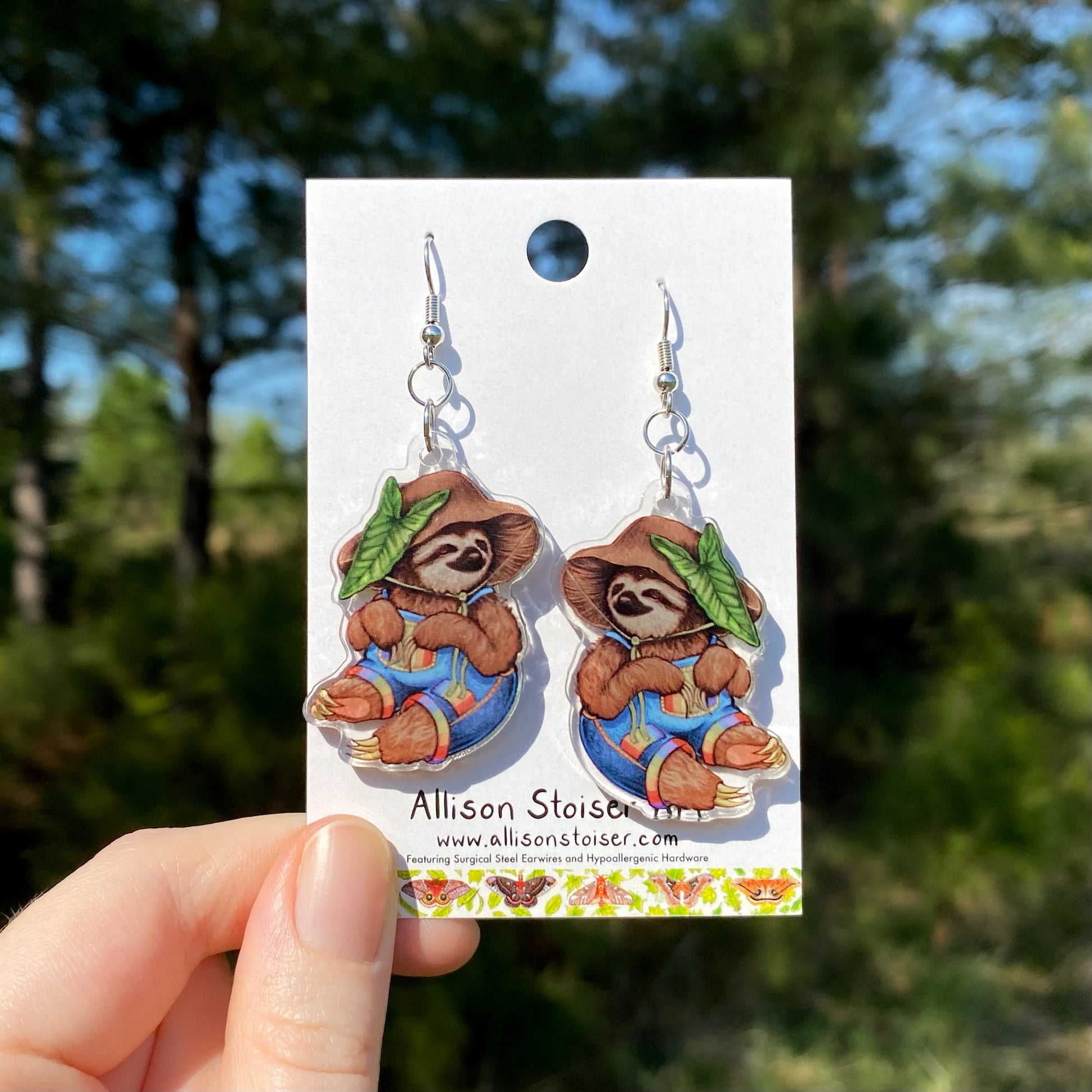 Sloth earrings on a card held by a hand with a blurred natural background