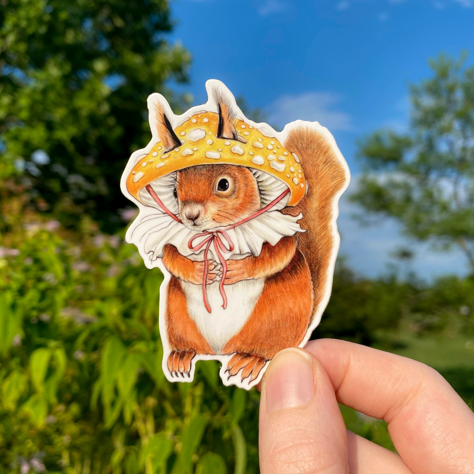 A hand holding a sticker of a red squirrel, wearing a yellow patches mushroom hat.