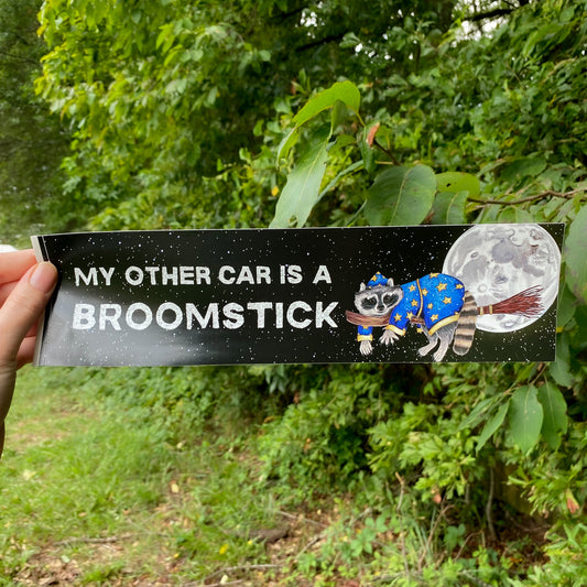 A bumper sticker featuring a broomstick-riding raccoon in front of a night sky. The text reads "My Other Car is a Broomstick."