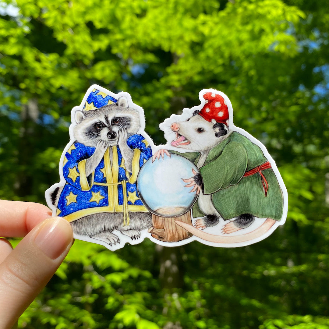 A hand holding a sticker featuring an illustration of a wizard raccoon and fortuneteller opossum