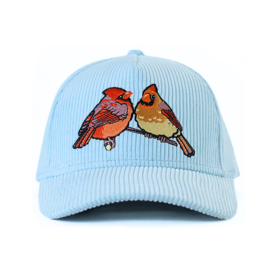 The front of a pale blue corduroy baseball cap with an embroidered pair of northern cardinals on a white background.