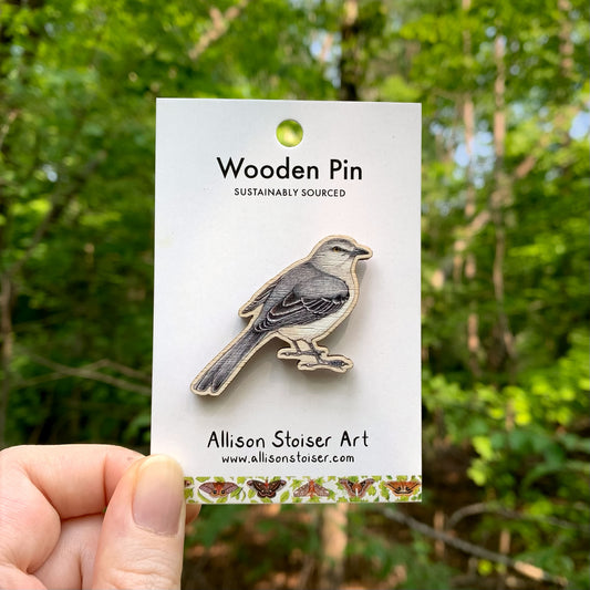 Mockingbird wooden pin on a card with 'Allison Stoiser Art' branding, held against a forest background.