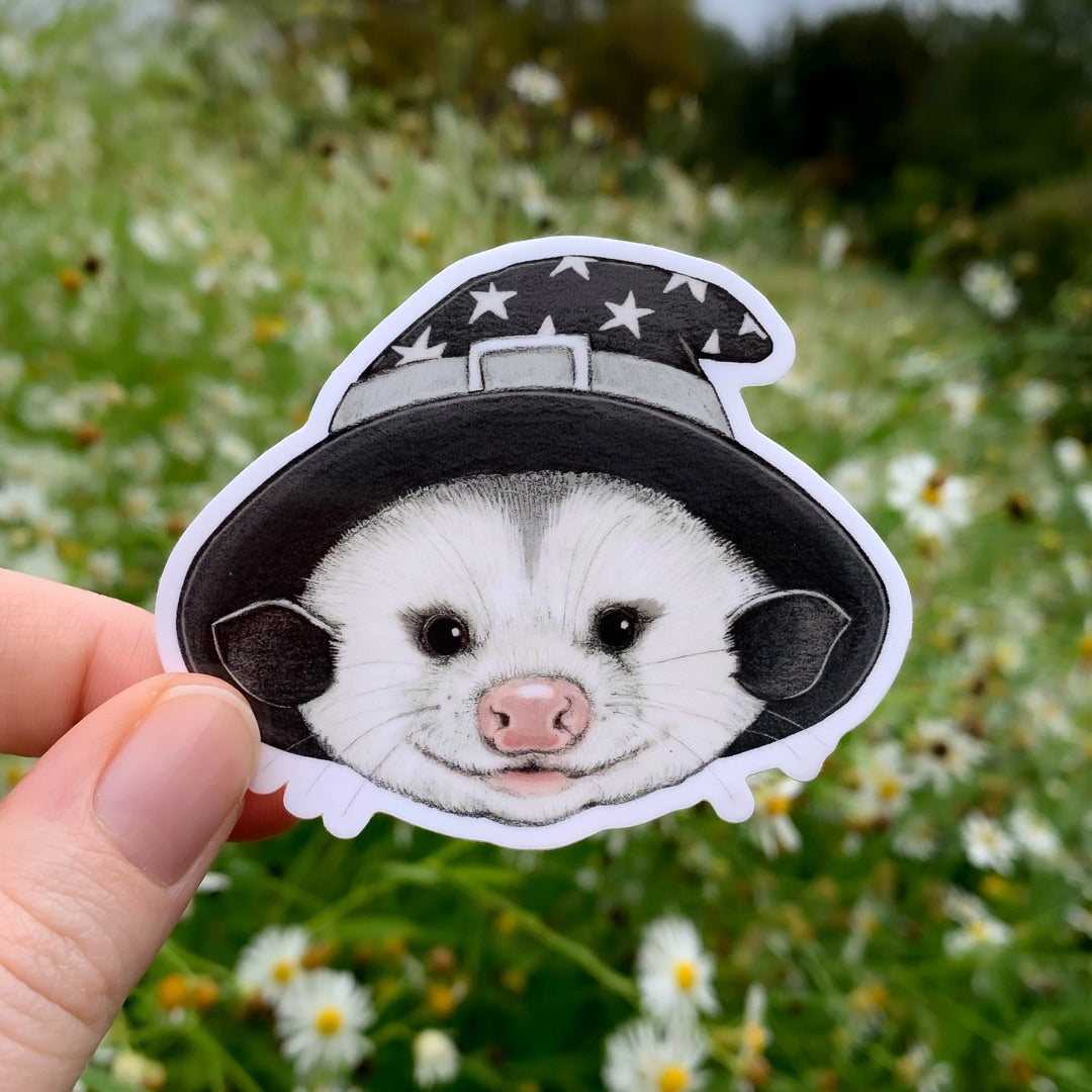 A hand holding a sticker of an opossum wizard wearing a black wizard hat with silver stars.