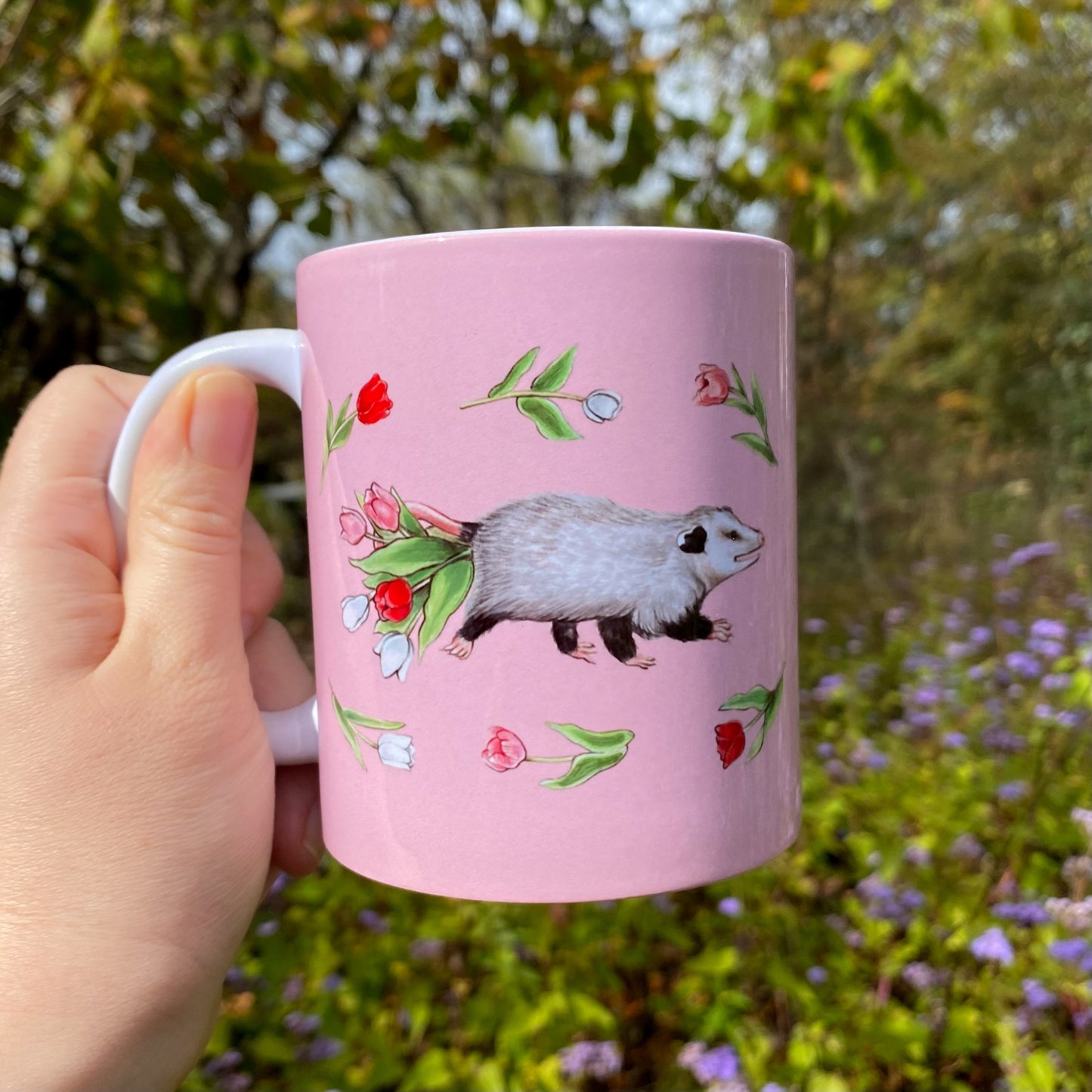 Pink mug with an illustration of an opossum and tulips held by a hand outdoors.