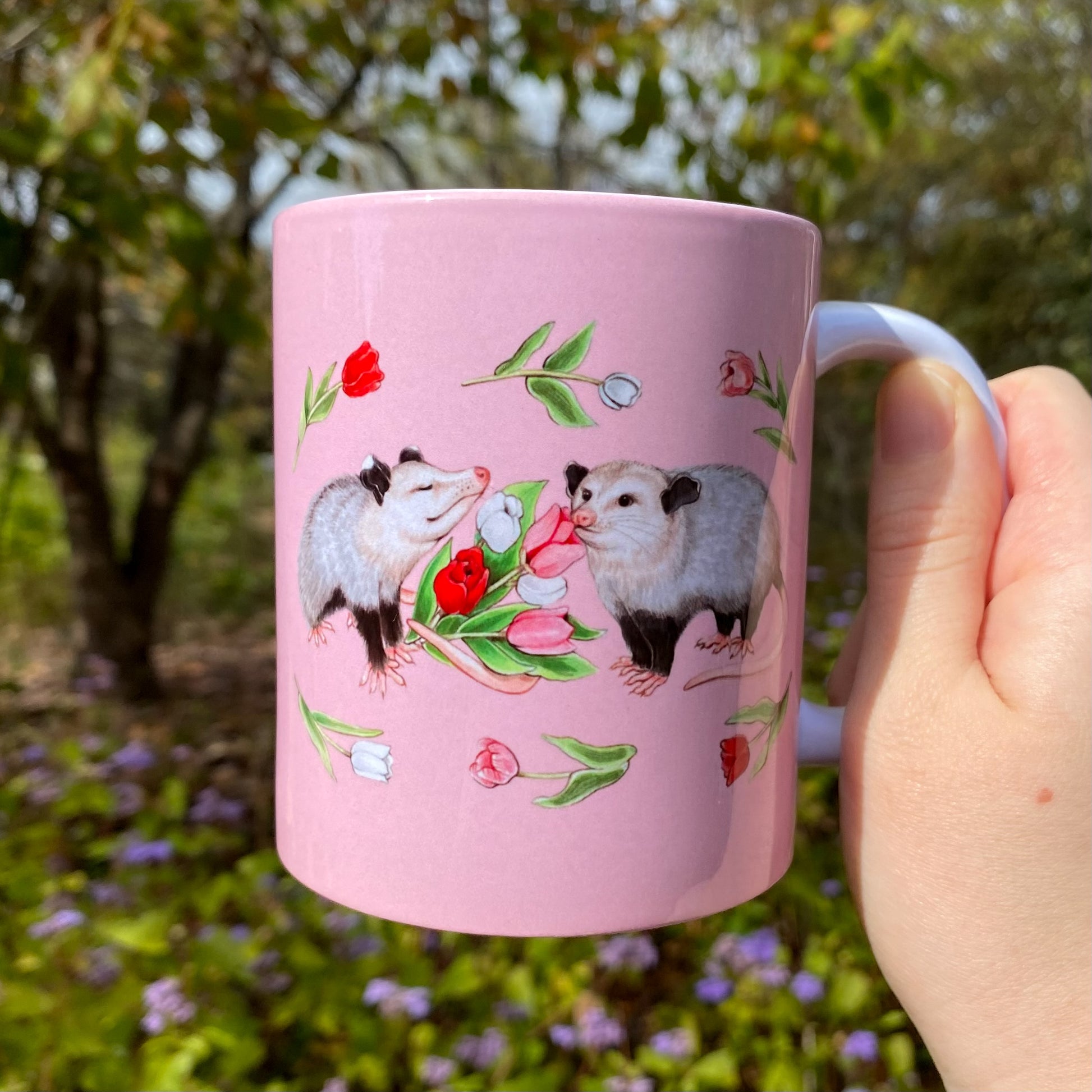 Pink mug with an opossum and tulip illustration held in a hand outdoors.
