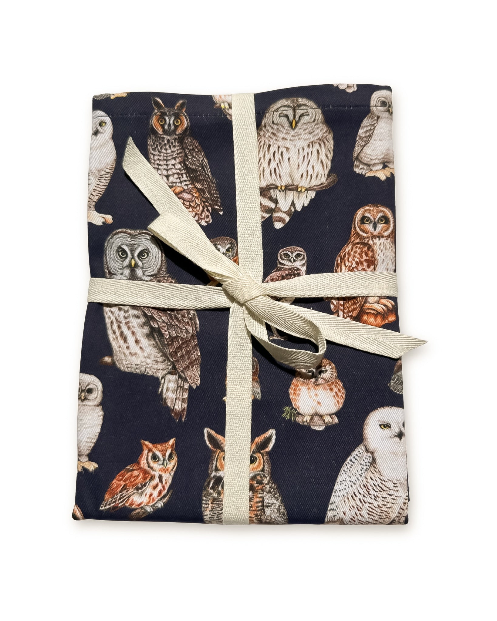 A tea towel featuring an all-over owl print on a dark navy background and tied with a cream ribbon, shown on a plain white background.
